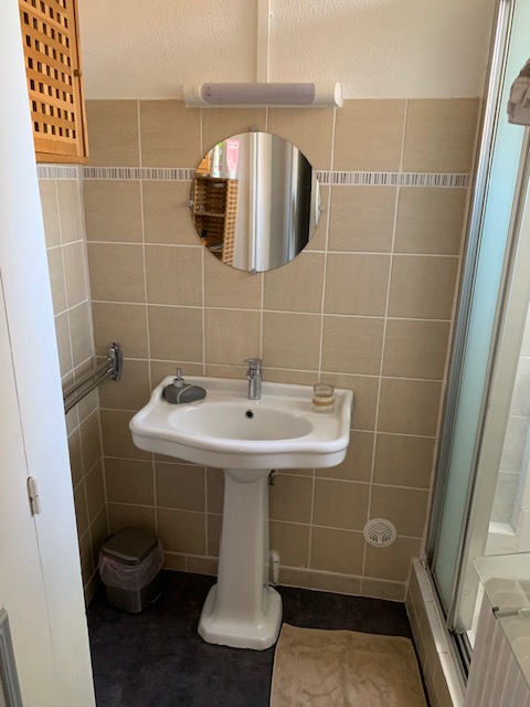 Salle d'eau carrelée beige avec lavabo sur colonne, miroir rond et cabine de douche
