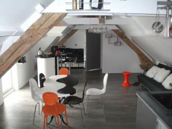 Loft sous charpente : table ronde, chaises orange et blanches, suspension en verre